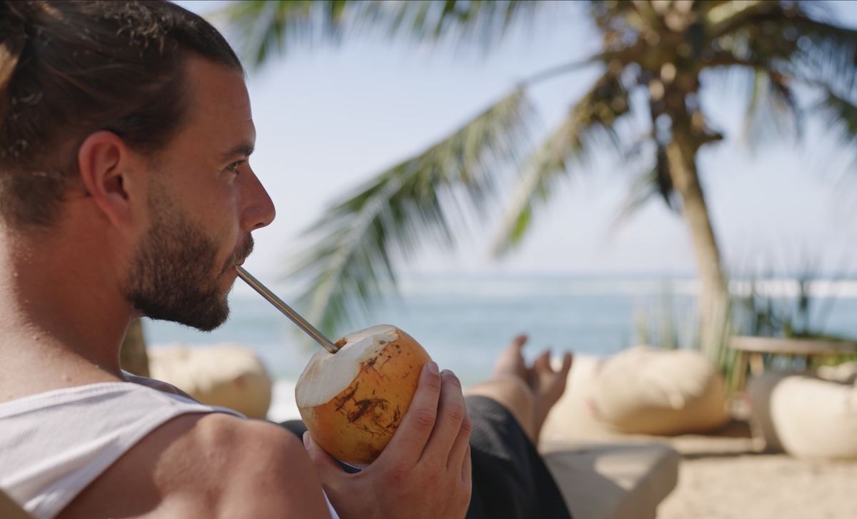 A guest relaxing with a coconut under a palm tree by the sea